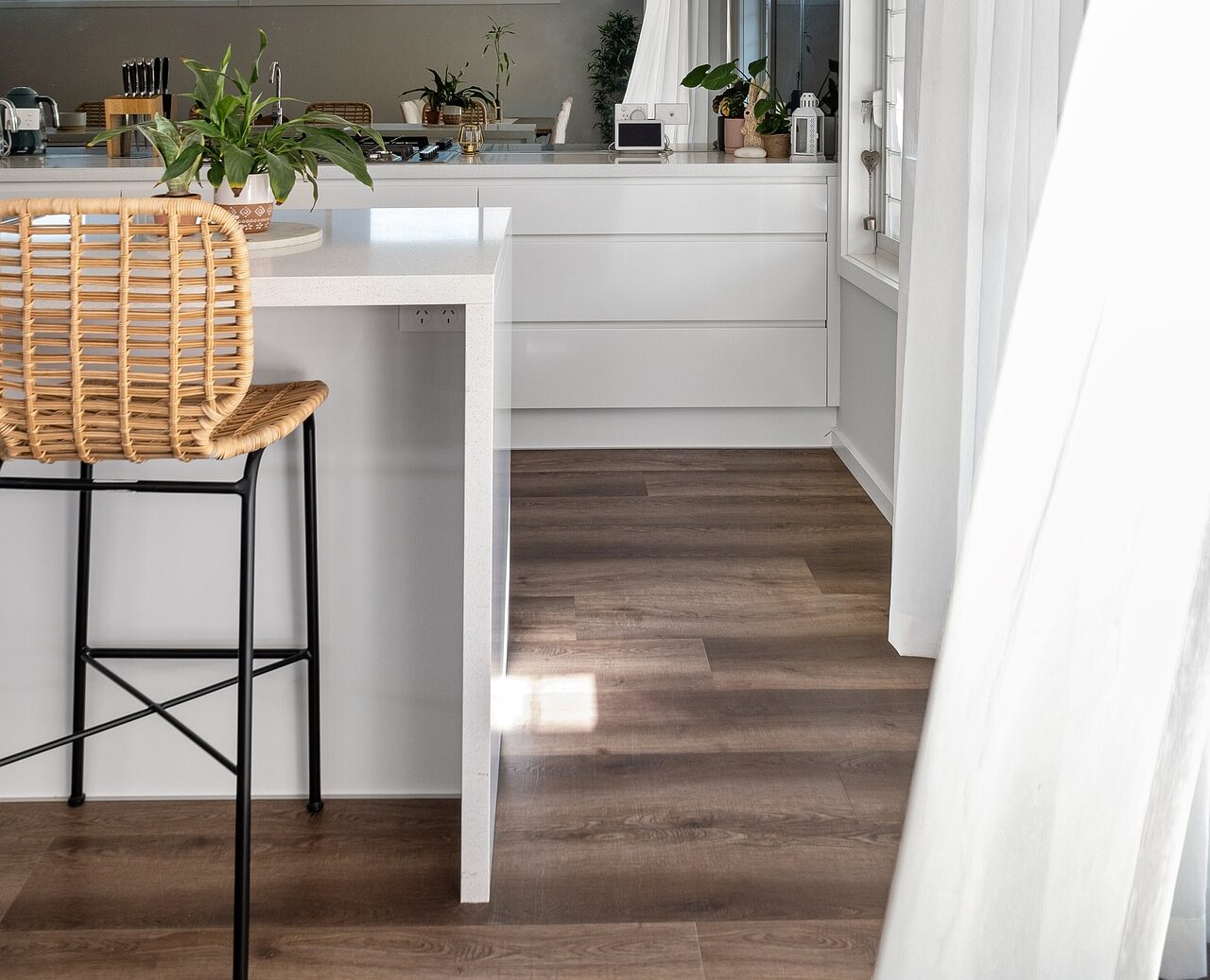 Vinyl-Designboden in dunkler Holzoptik in einer hellen Küche mit weißen Fronten und Barhocker aus Rattan