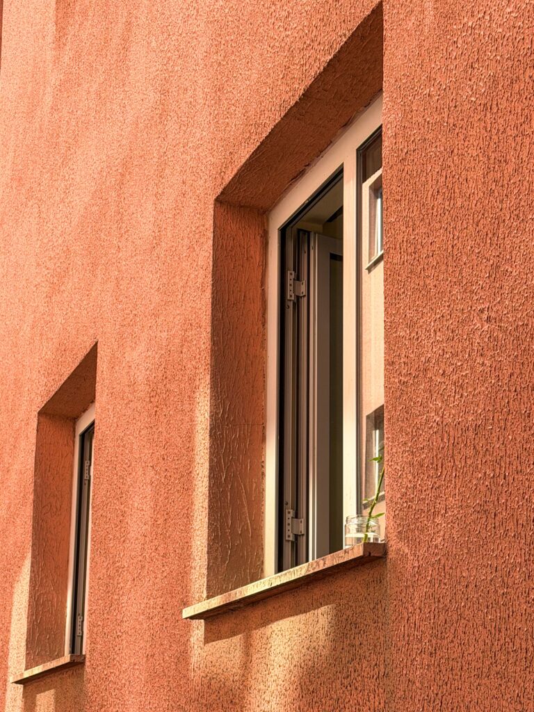 Fassade mit grob strukturiertem Putz in warmem Terrakotta-Ton und Fenstern mit tiefen Laibungen im Sonnenlicht