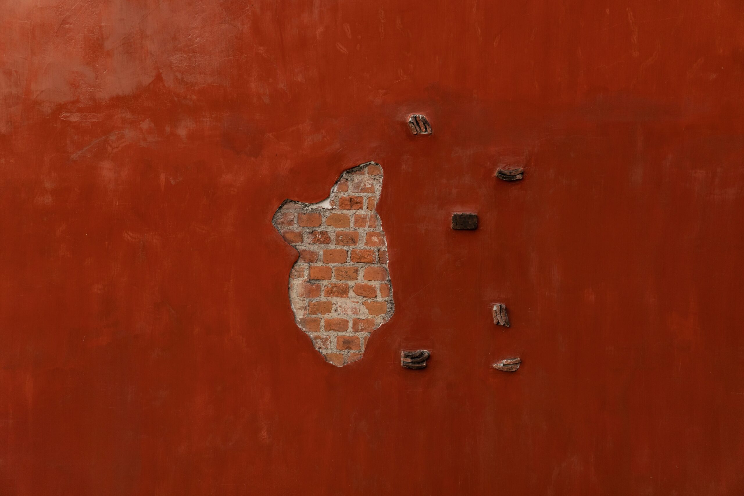 Rot gestrichene Putzwand mit einer Fehlstelle, durch die das Ziegelmauerwerk sichtbar ist, und alten Haken in der Wand