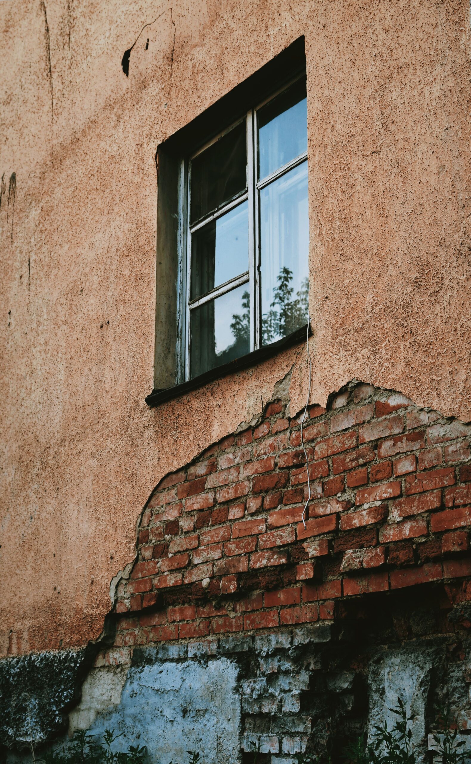 Sanierungsbedürftige Fassade, bei der großflächig abgeplatzter Putz unter einem Fenster das Ziegelmauerwerk freilegt