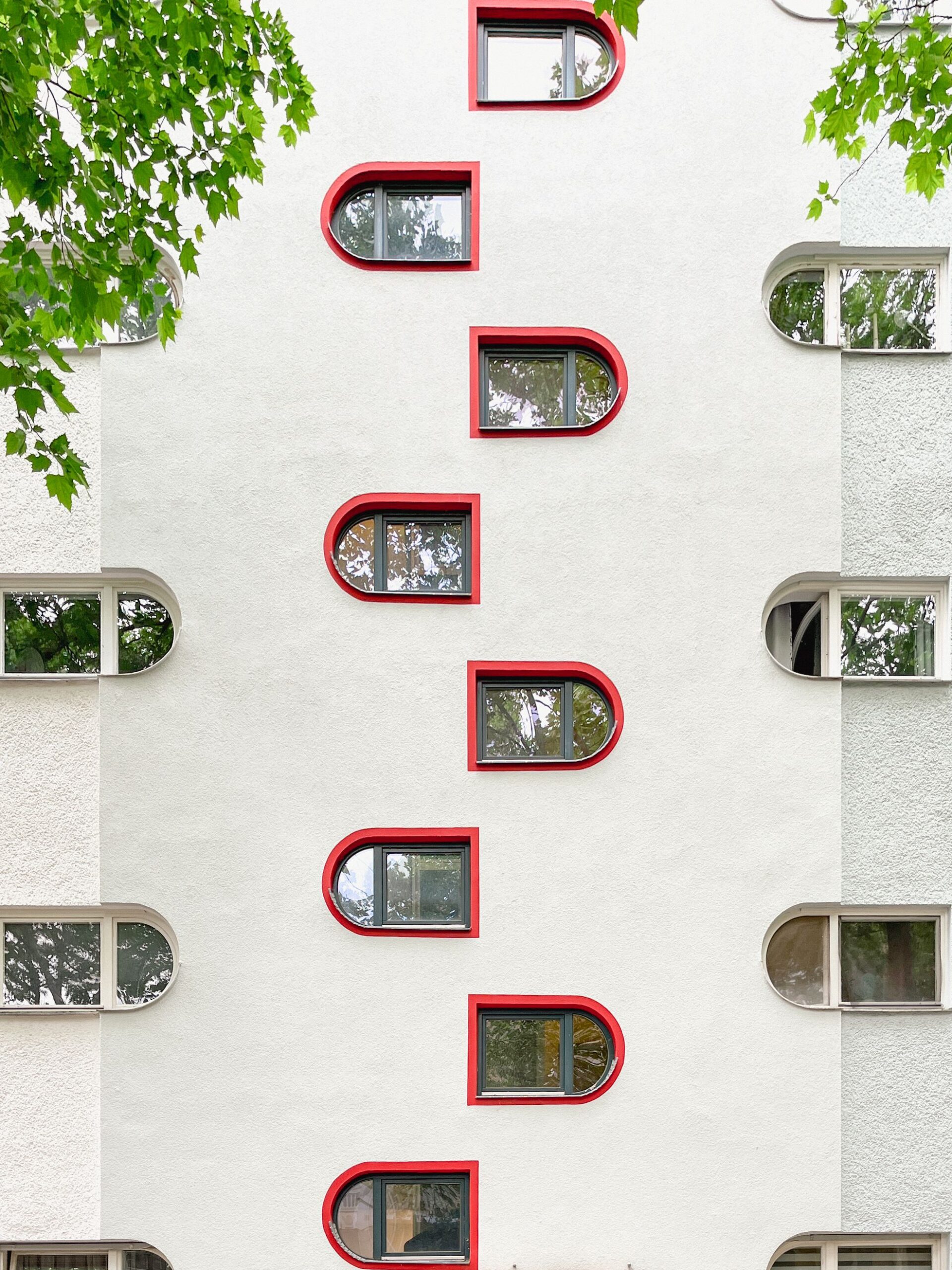 Weiße Putzfassade mit versetzt angeordneten, abgerundeten Fenstern, deren Laibungen rot abgesetzt sind